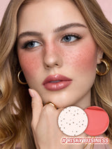 Close-up of a woman with freckles wearing a glossy peachy-pink lip color and rosy blush, with two circular makeup swatches at the bottom showing the shade and texture. The text '#RISKY BUSINESS' appears in the lower right corner.