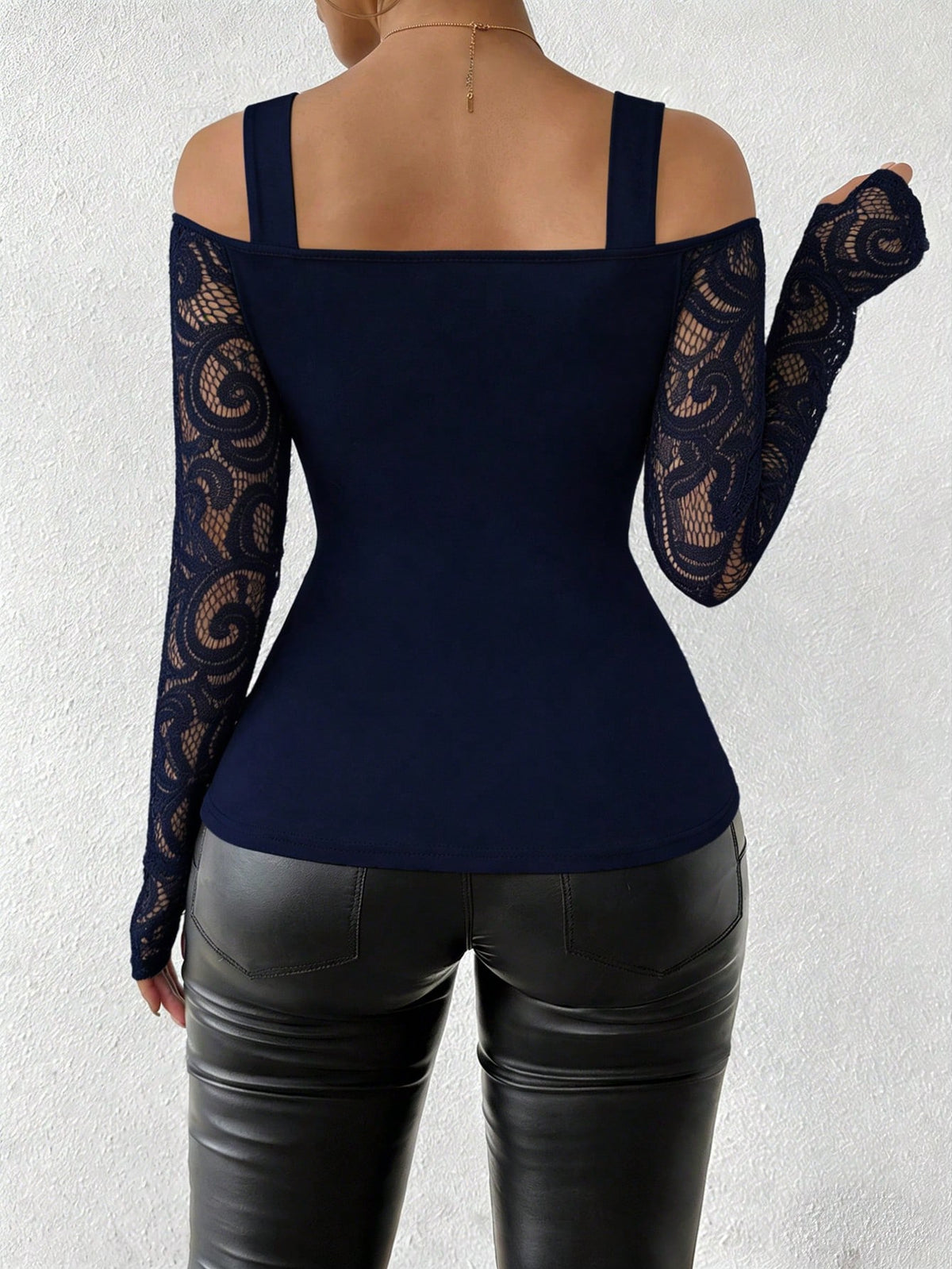 A woman shown from the back wearing a navy blue cold-shoulder top with long lace sleeves and black faux-leather pants, against a light textured wall.