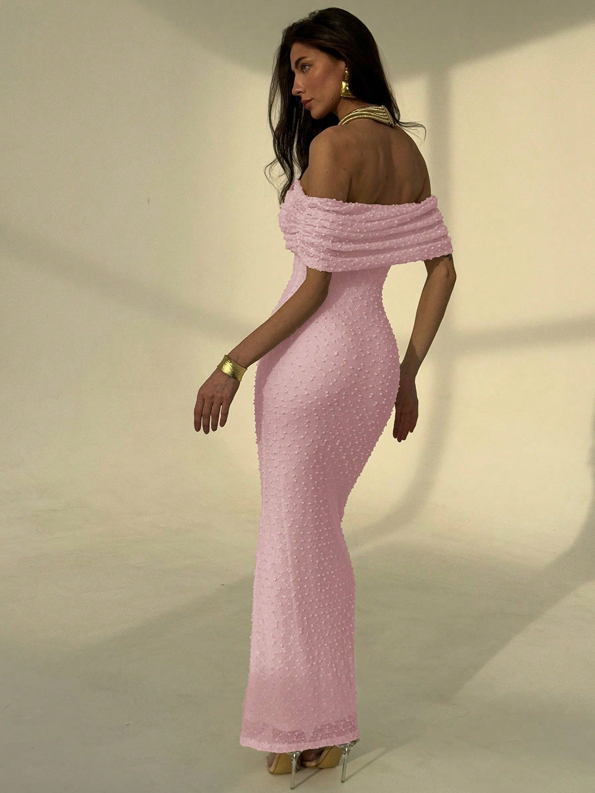 A woman modeling a light pink off-the-shoulder maxi dress with a textured, dotted fabric and a fitted silhouette, shown from a side-back angle against a warm beige studio background.