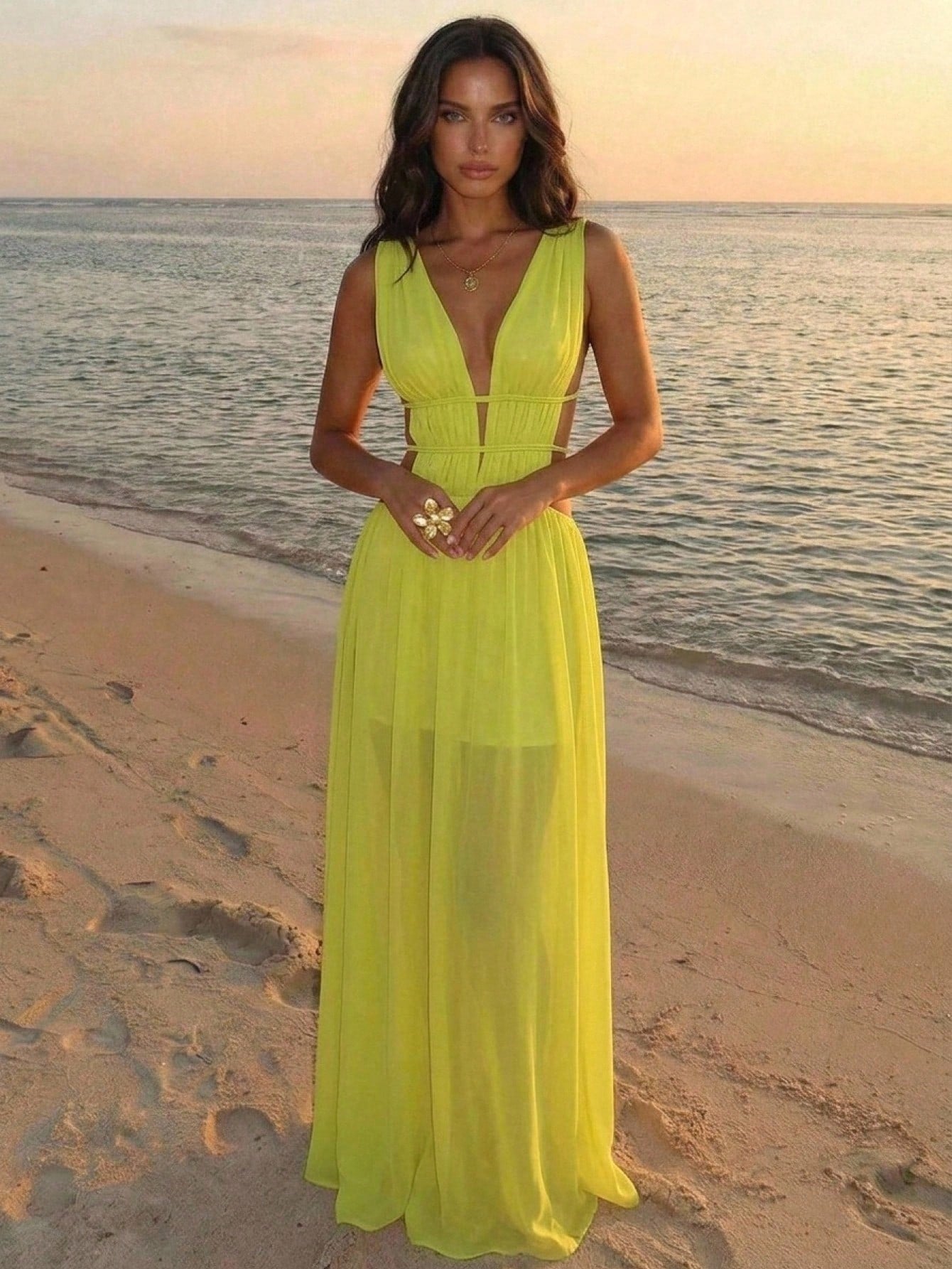 A woman stands on a sandy beach at sunset wearing a bright neon yellow sleeveless maxi dress with a deep V-neckline and a flowing chiffon-like skirt.