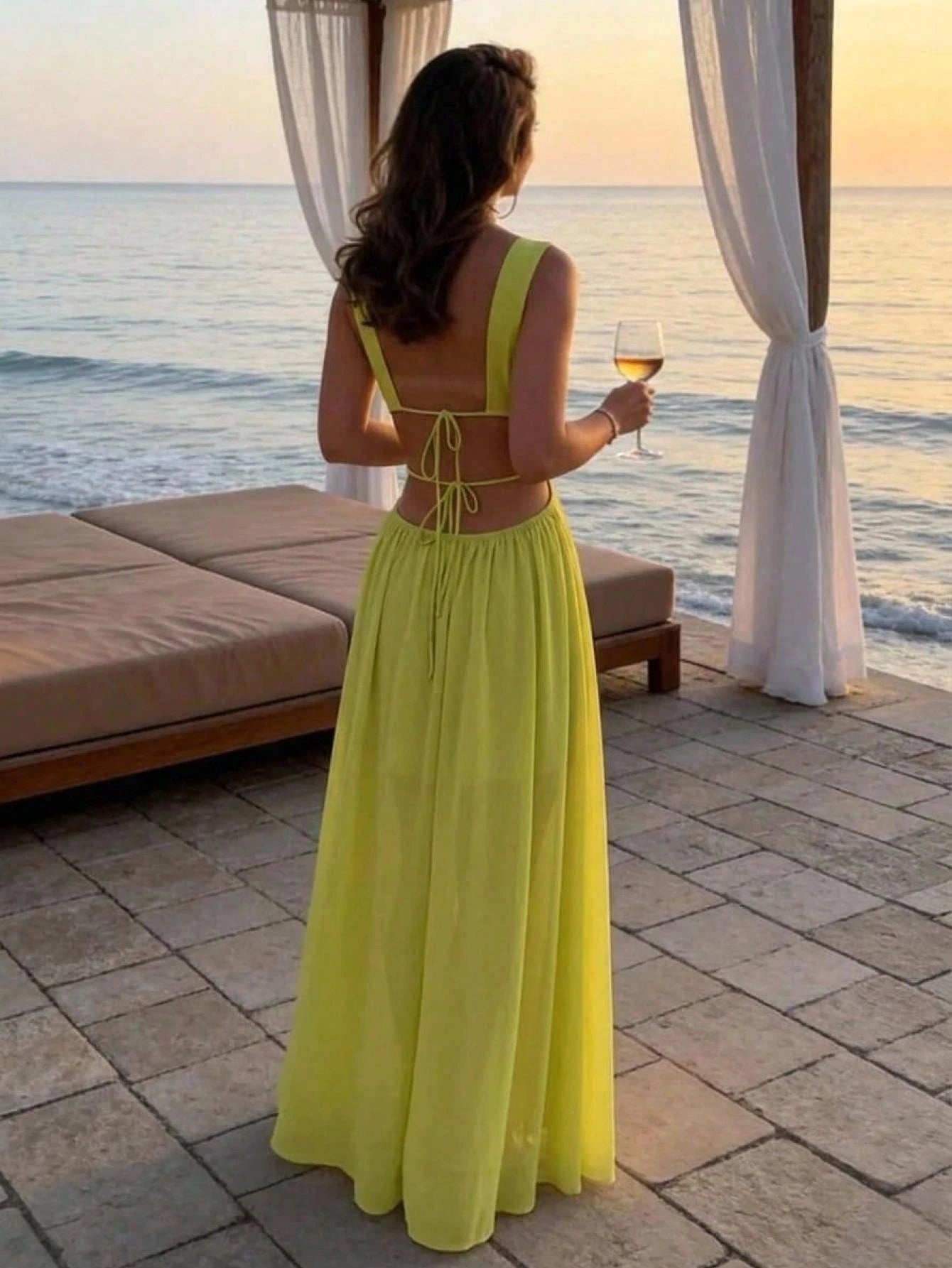 A woman stands on a beachside patio at sunset wearing a bright yellow-green sleeveless maxi dress with a tie-back back and holding a glass of white wine.