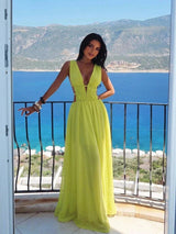 A woman wearing a bright yellow-green sleeveless maxi dress with a deep V-neckline stands on a balcony overlooking a scenic blue sea and mountains.
