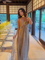 A woman wearing a strapless pleated champagne-gold maxi dress with a gold choker and statement necklace, standing in a modern Japanese-style room with shoji screens and a long dining table.