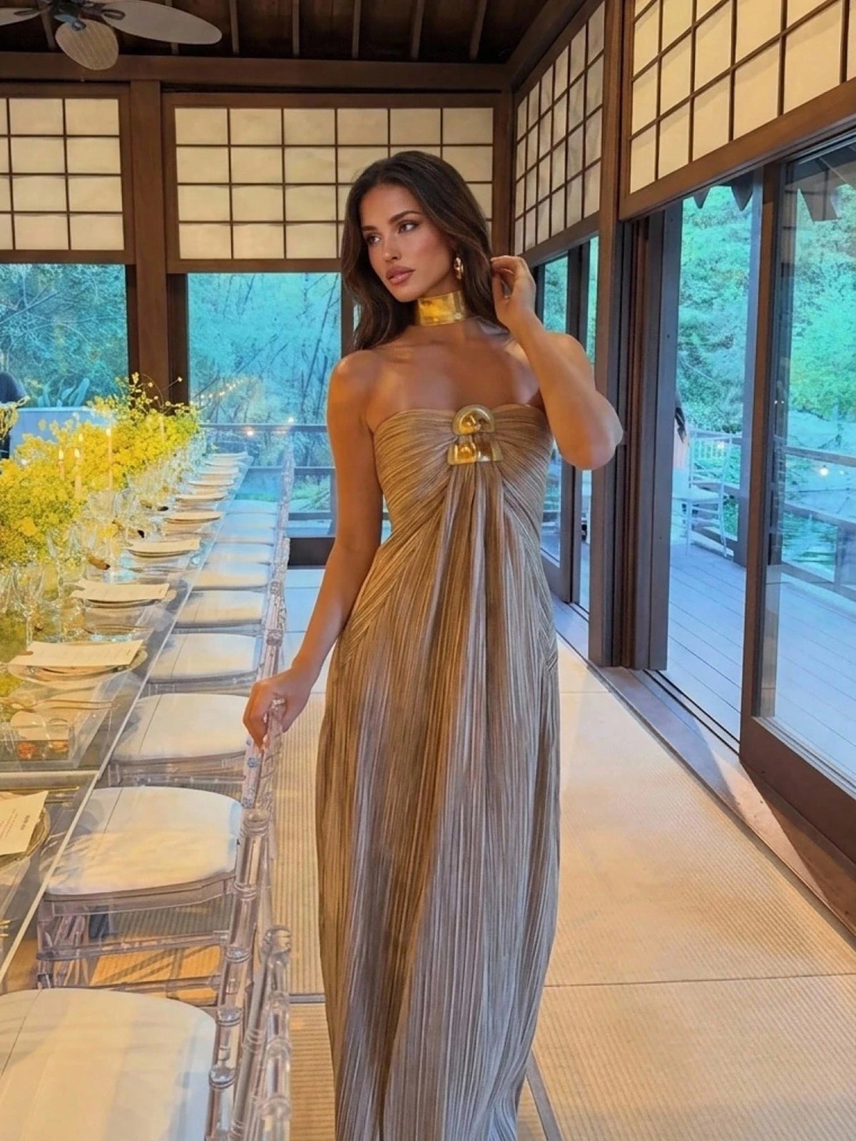 A woman wearing a strapless pleated champagne-gold maxi dress with a gold choker and statement necklace, standing in a modern Japanese-style room with shoji screens and a long dining table.