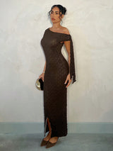 A woman models a textured, off-the-shoulder dark brown maxi dress with a side slit, paired with brown pointed-toe heels and holding a small metallic clutch, standing against a textured off-white wall.