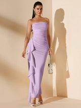 A woman models a strapless lavender maxi dress with a ruched bodice and dramatic layered ruffle draping down the left side, styled with metallic silver sandals and a small silver handbag against a beige wall with a strong shadow.
