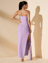 A woman stands with her back to the camera wearing a strapless lavender maxi dress with a high thigh slit, photographed in a studio with warm beige walls and a strong cast shadow.