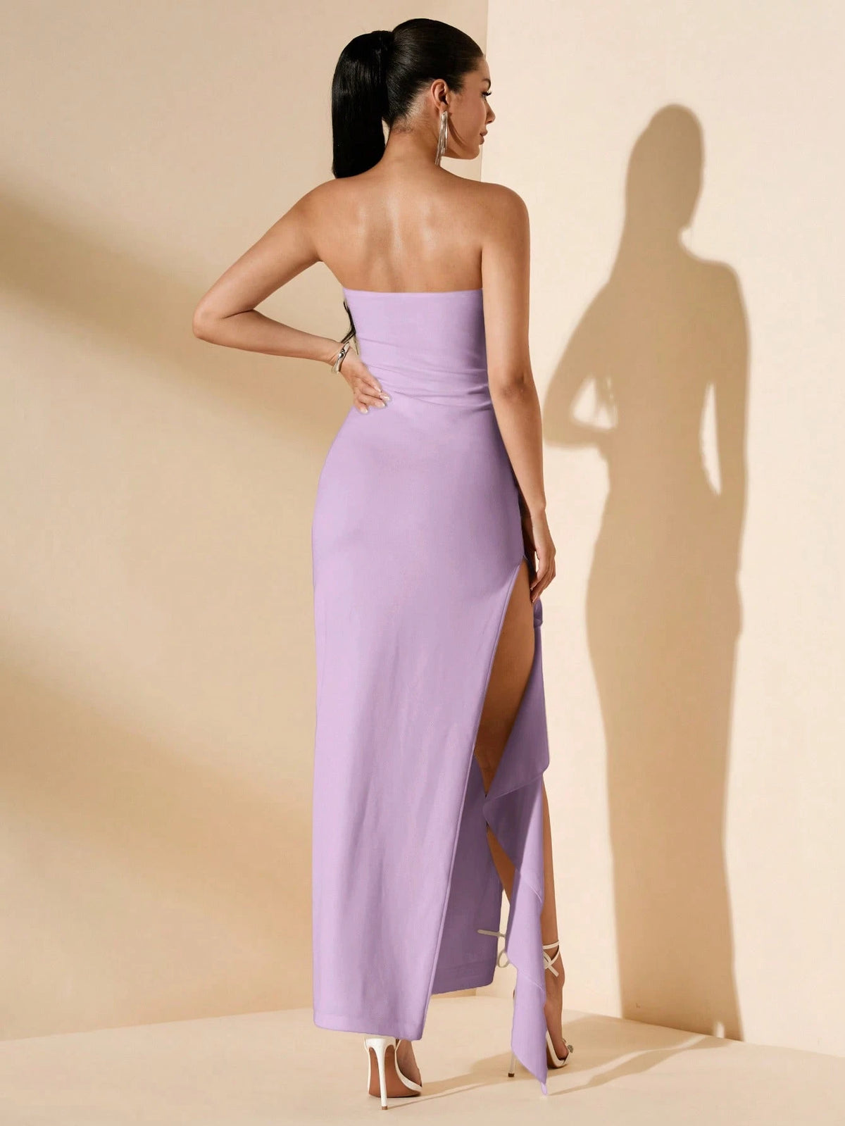 A woman stands with her back to the camera wearing a strapless lavender maxi dress with a high thigh slit, photographed in a studio with warm beige walls and a strong cast shadow.