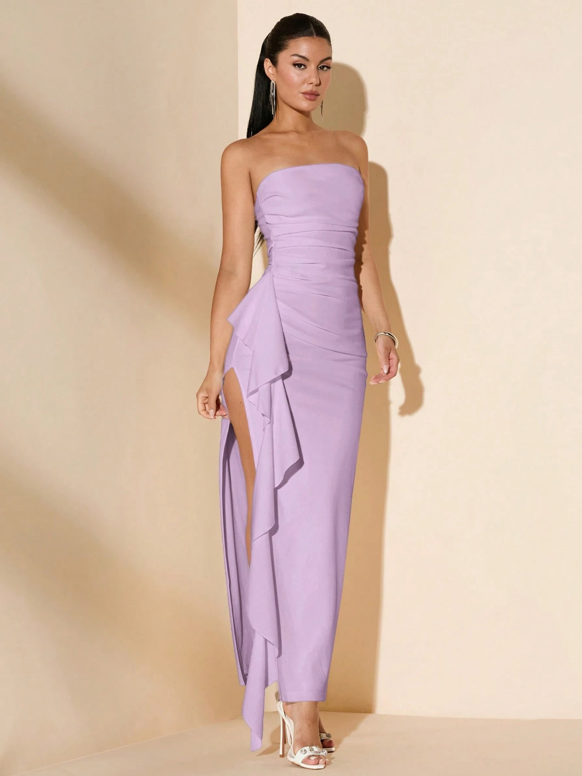 A woman modeling a strapless lavender maxi dress with a ruched bodice, side slit, and layered ruffle detail, standing against a warm beige studio backdrop.
