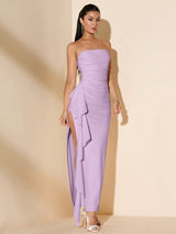 A woman modeling a strapless lavender maxi dress with a ruched bodice, side slit, and layered ruffle detail, standing against a warm beige studio backdrop.