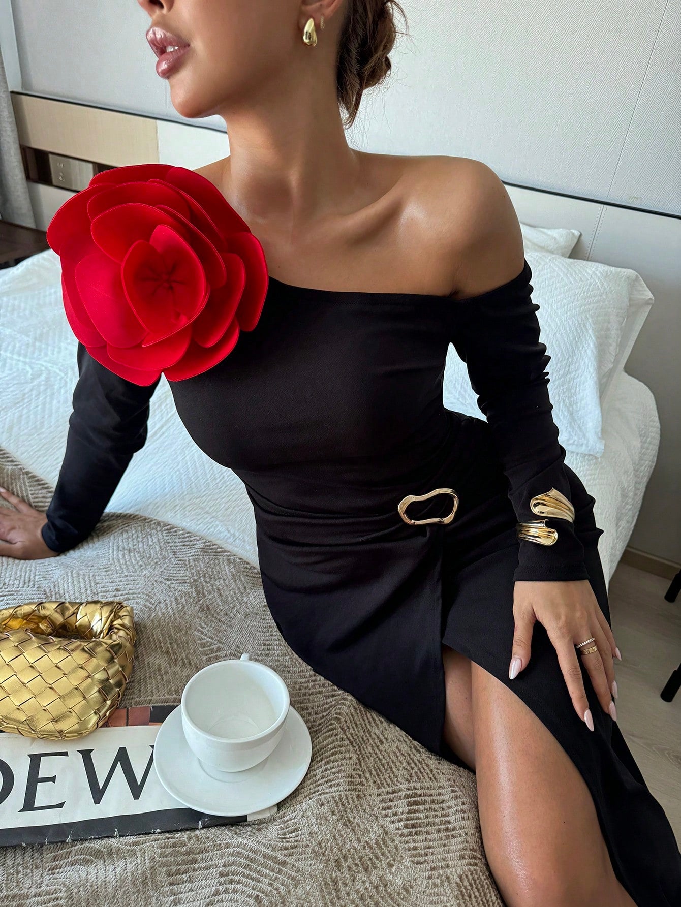 Woman in a black off-shoulder dress with a red flower and gold accessories on a bed.