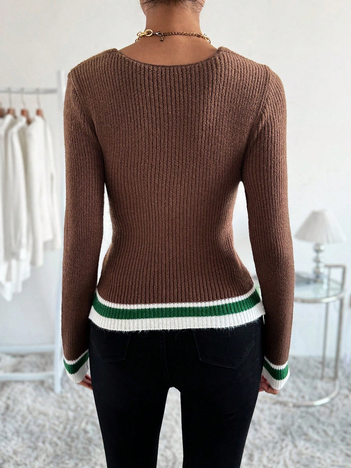 A woman viewed from the back wearing a fitted brown ribbed knit sweater with green and white striped trim at the hem and cuffs, paired with black pants in a bright indoor room.