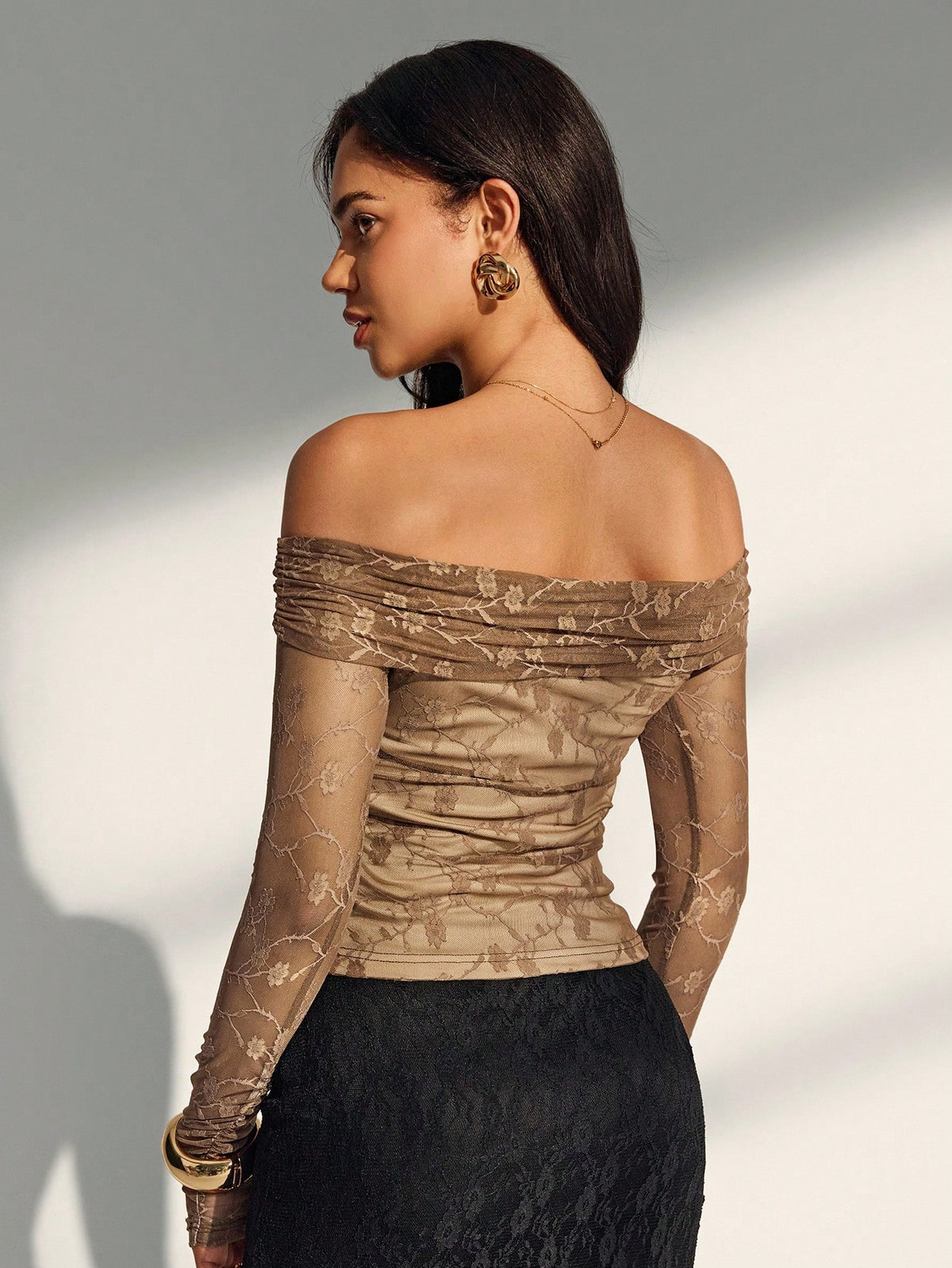 A woman shown from the back wearing an off-the-shoulder ruched top with sheer patterned sleeves in a taupe-beige tone, paired with a black skirt, against a softly lit gray background.