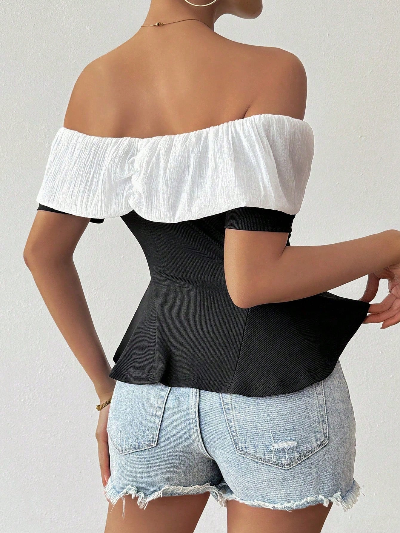 Person wearing a white off-shoulder top over a black peplum top with denim shorts on a light gray background