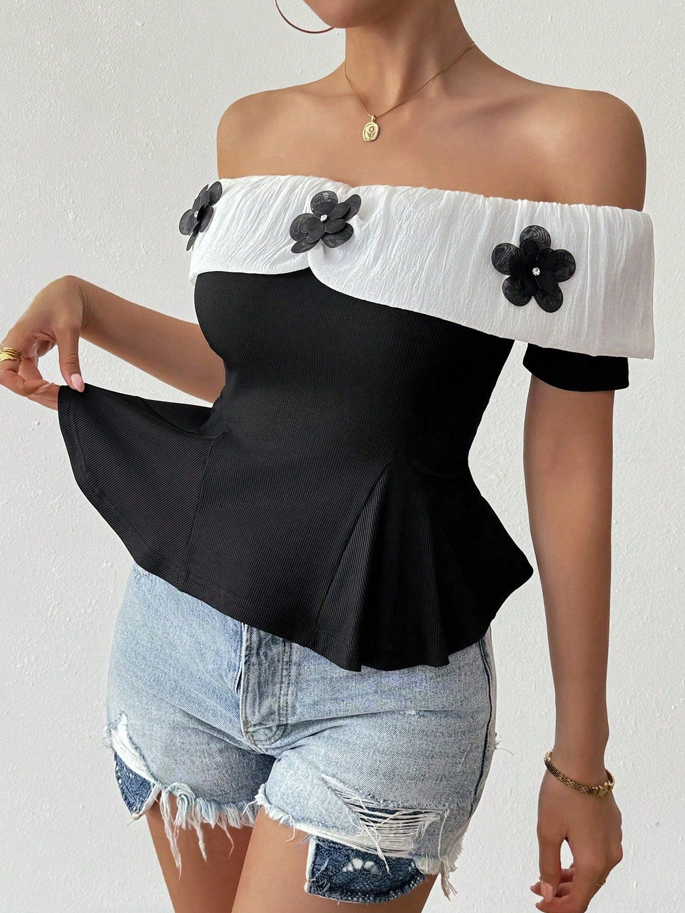 Person wearing a black and white off-shoulder top with floral details and denim shorts on a plain background