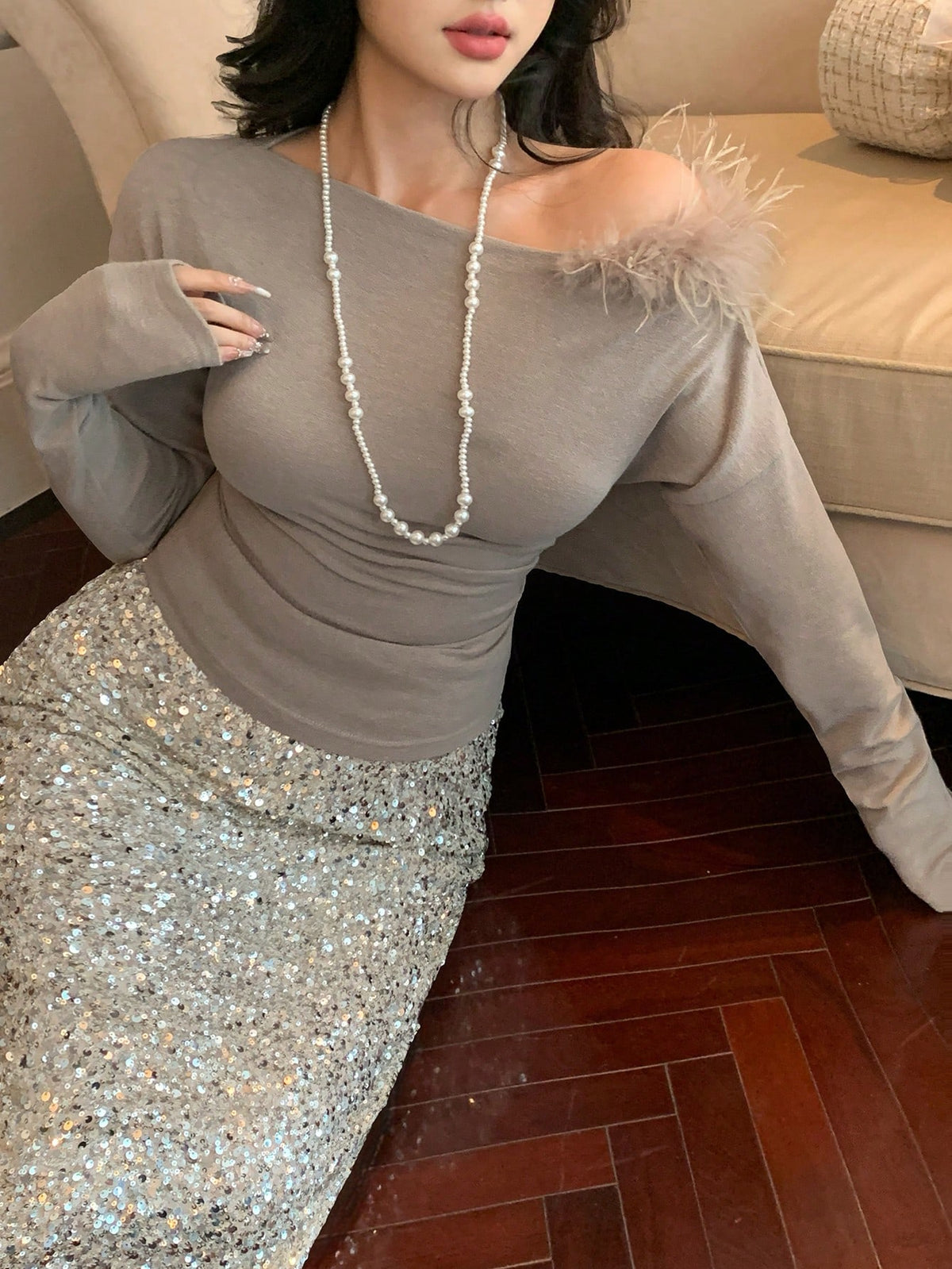 A woman wearing a taupe-gray long-sleeve top with feather trim at the shoulder and a sparkling silver sequined skirt, posed in an indoor setting with a wooden floor and sofa in the background.