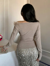 A woman shown from behind wearing a taupe long-sleeve top with feathered shoulder detailing and a silver sequined skirt, standing indoors next to a table.
