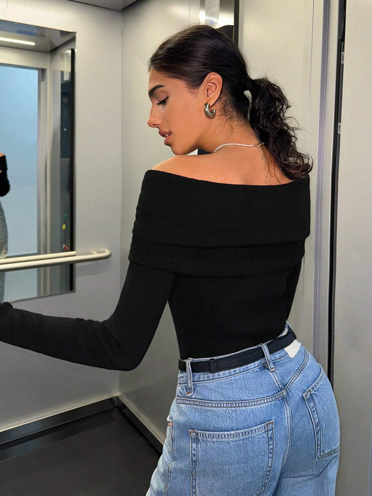 A woman wearing a black off-the-shoulder long-sleeve top and light blue jeans, standing inside a train or subway car next to a window.