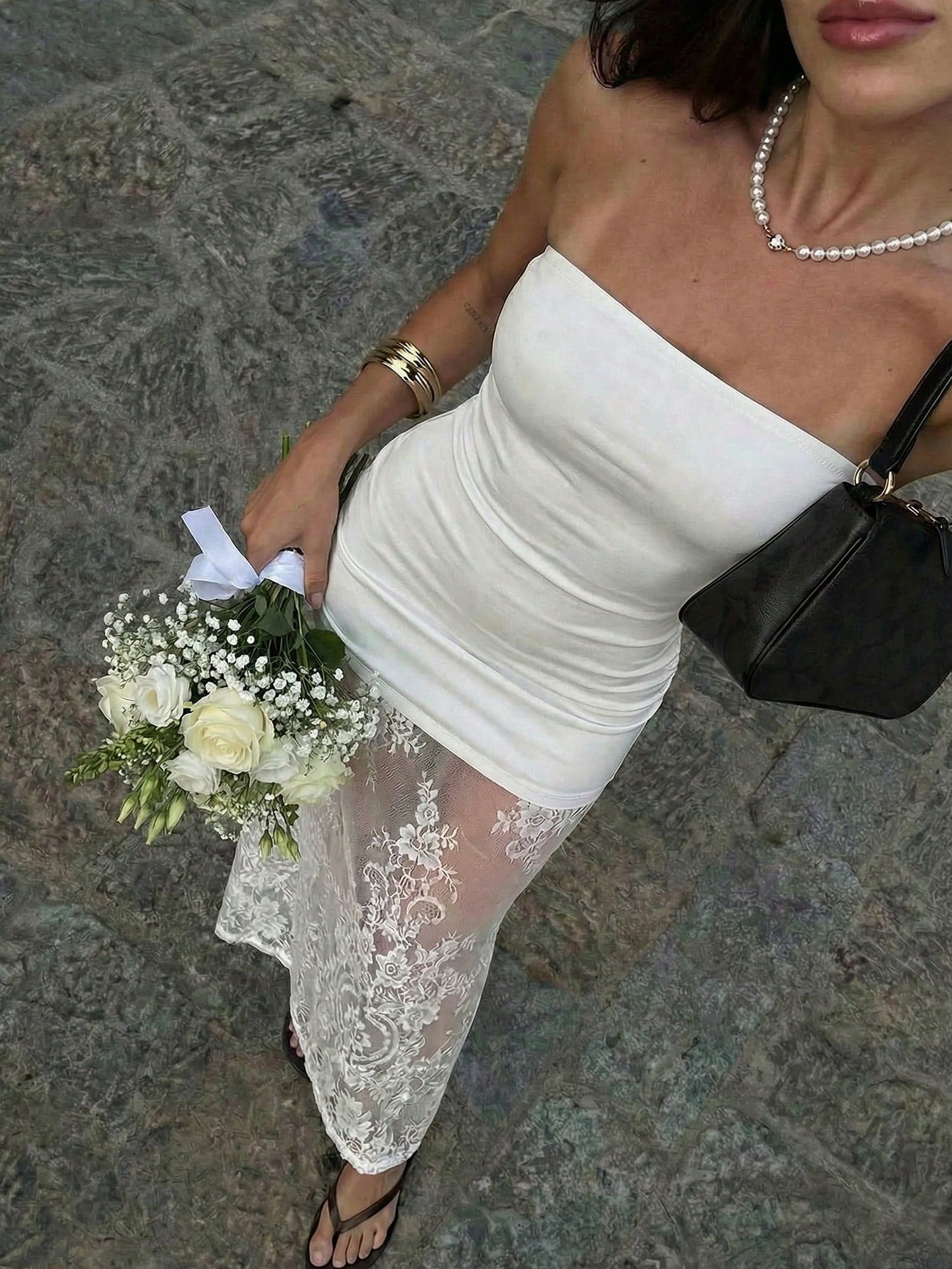 Woman in a white off-shoulder dress with lace detailing holding a bouquet on a stone pavement.
