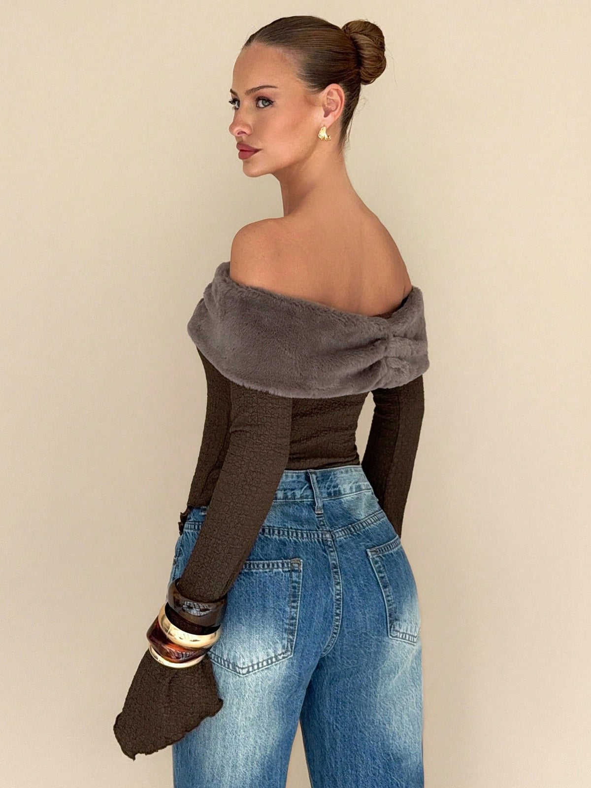A woman shown from the back wearing a dark brown off-the-shoulder long-sleeve top with a wide fuzzy taupe-gray collar, paired with blue jeans and stacked bracelets, against a plain beige background.