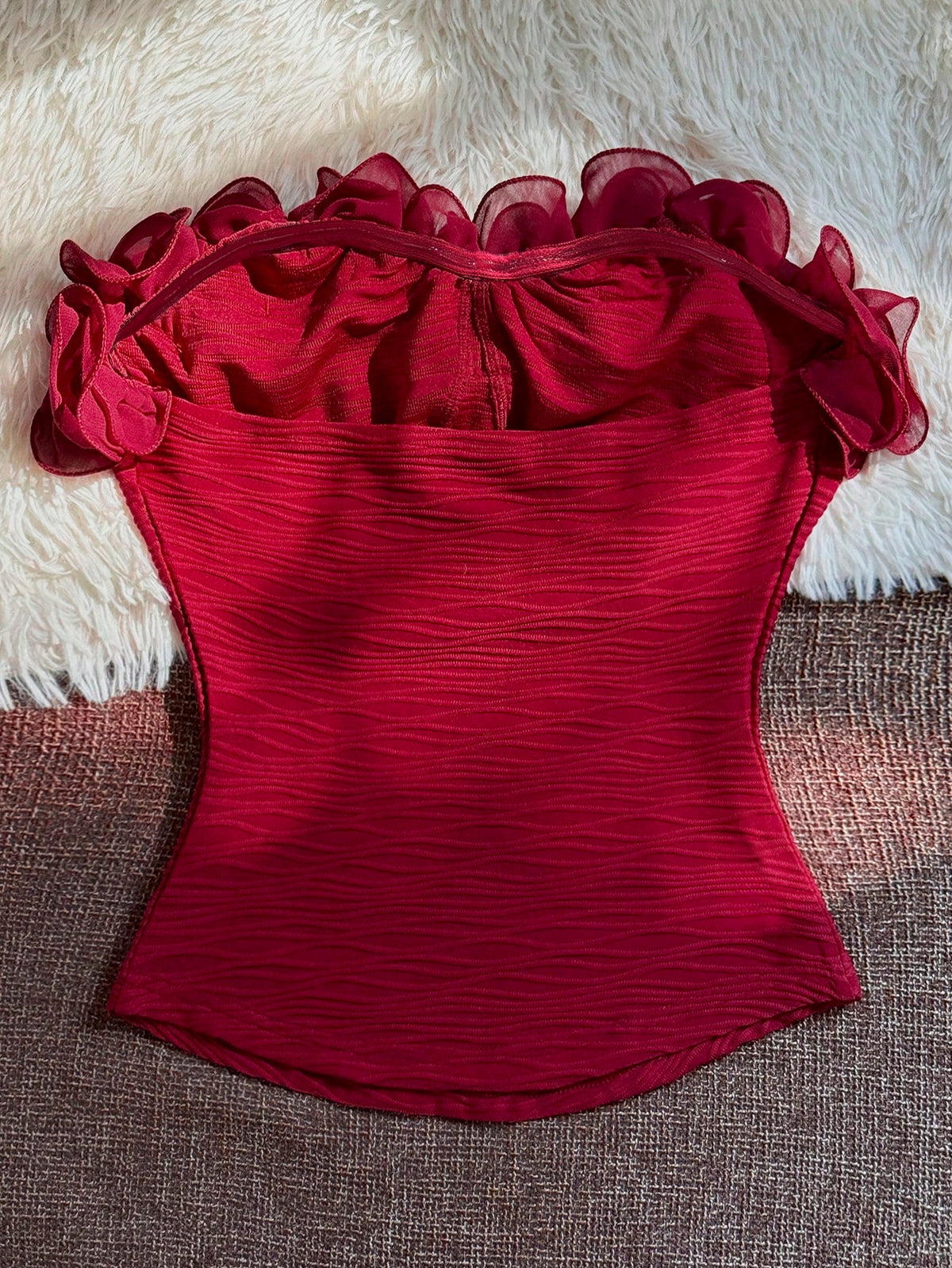 A red strapless corset-style top with ruffled sheer shoulder details and a textured crinkled fabric, laid flat on a brown woven surface with a white faux-fur backdrop.