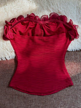 A red strapless corset-style top with ruffled sheer shoulder details and a textured crinkled fabric, laid flat on a brown woven surface with a white faux-fur backdrop.
