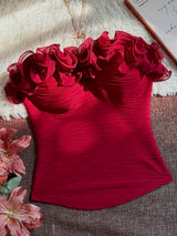 A deep red strapless corset-style top with layered ruffle details across the bust, laid flat on a textured brown surface with a white faux-fur backdrop, pink flowers, and a notebook corner visible.