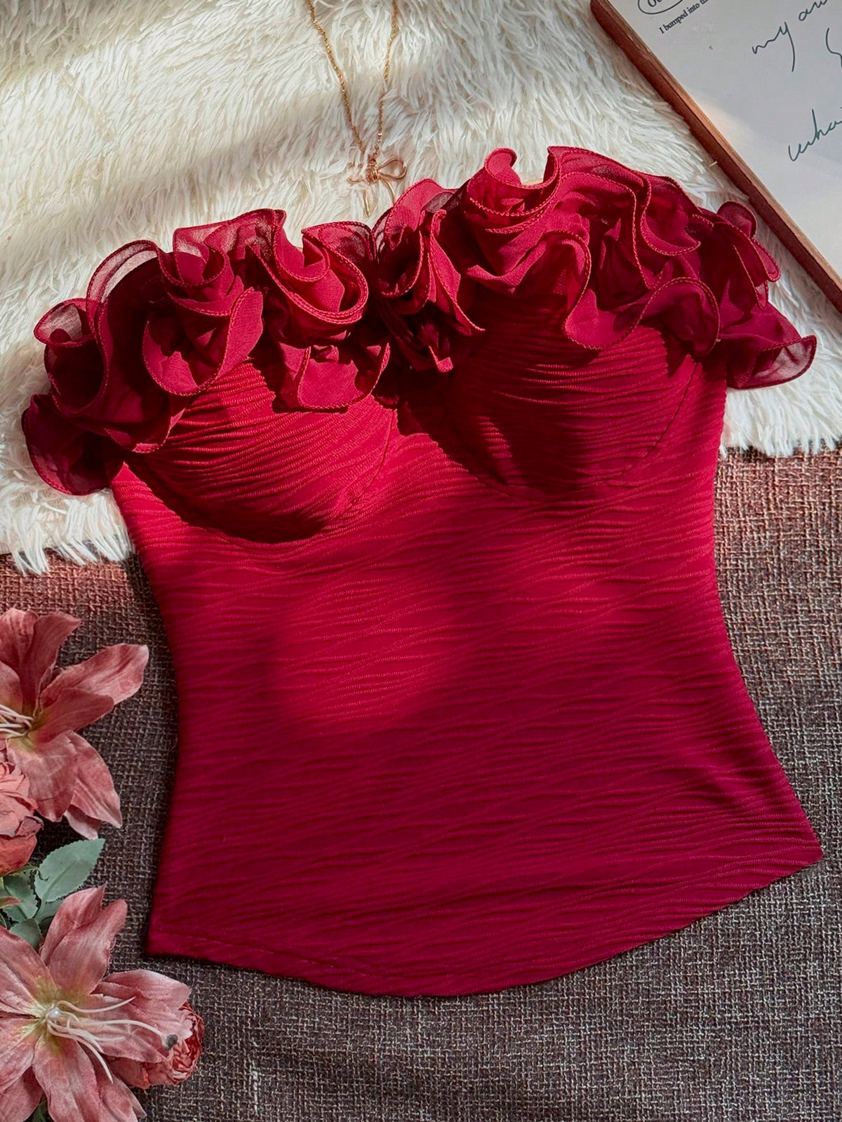 A deep red strapless corset-style top with layered ruffle details across the bust, laid flat on a textured brown surface with a white faux-fur backdrop, pink flowers, and a notebook corner visible.