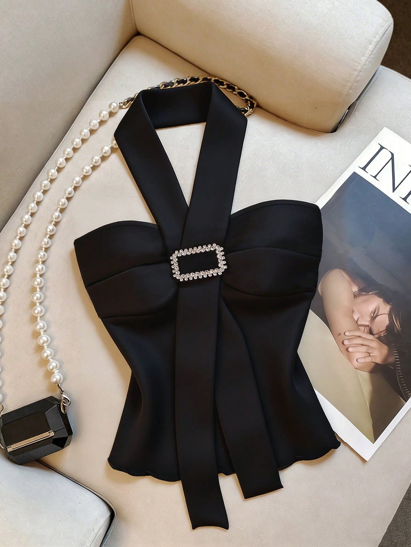 A black satin-like halter top with a large front bow and a rhinestone-embellished rectangular buckle, styled on a cream sofa with a pearl bag and magazine nearby.