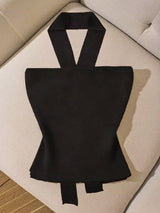 A black halter-style top with a structured, hourglass-shaped bodice and wide straps, laid flat on a light beige upholstered chair.