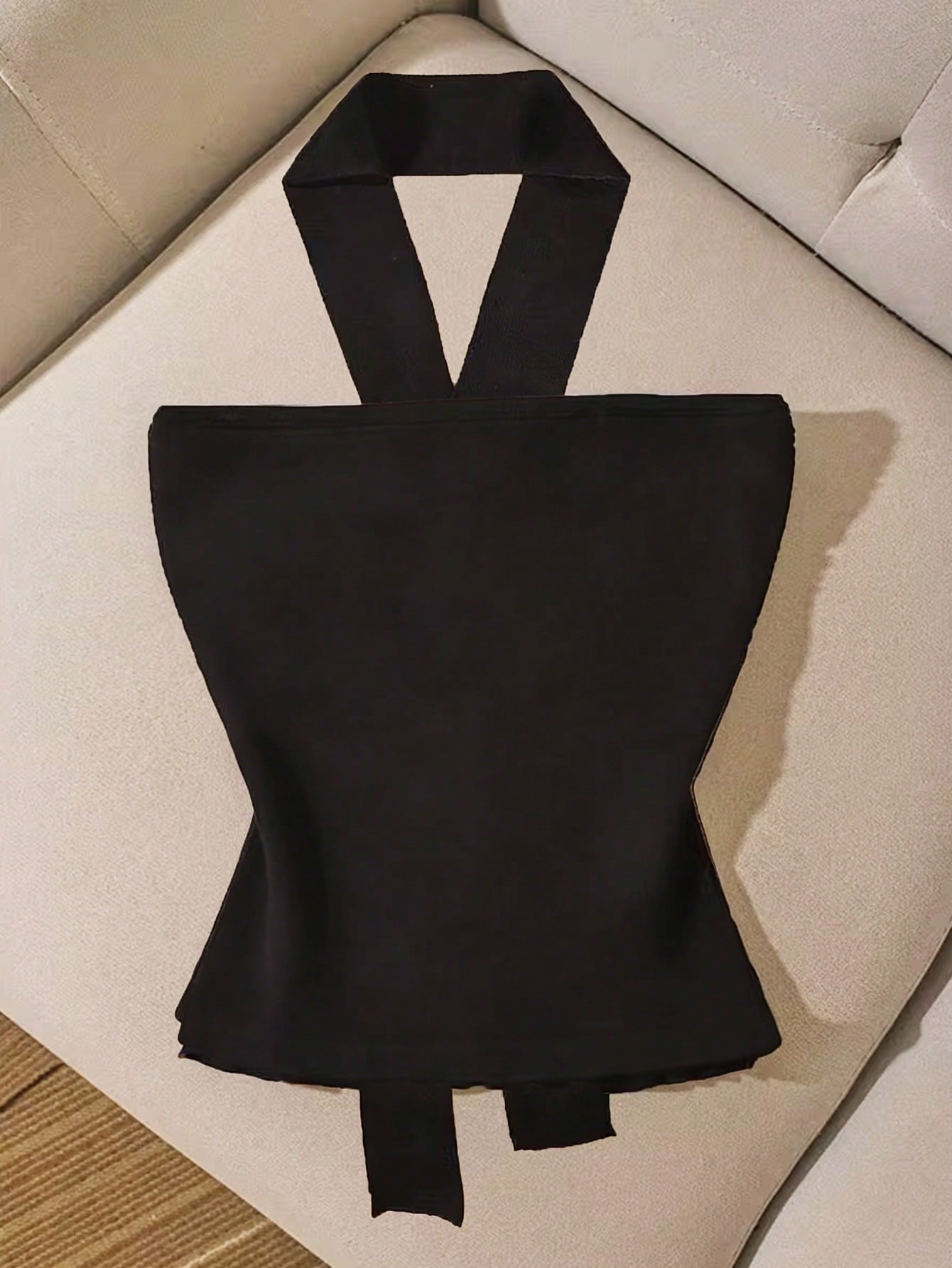A black halter-style top with a structured, hourglass-shaped bodice and wide straps, laid flat on a light beige upholstered chair.