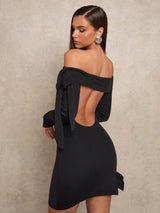 A woman modeling a black off-the-shoulder mini dress with a large open back, long sleeves, and a bow detail at the hem, photographed against a textured beige studio backdrop.