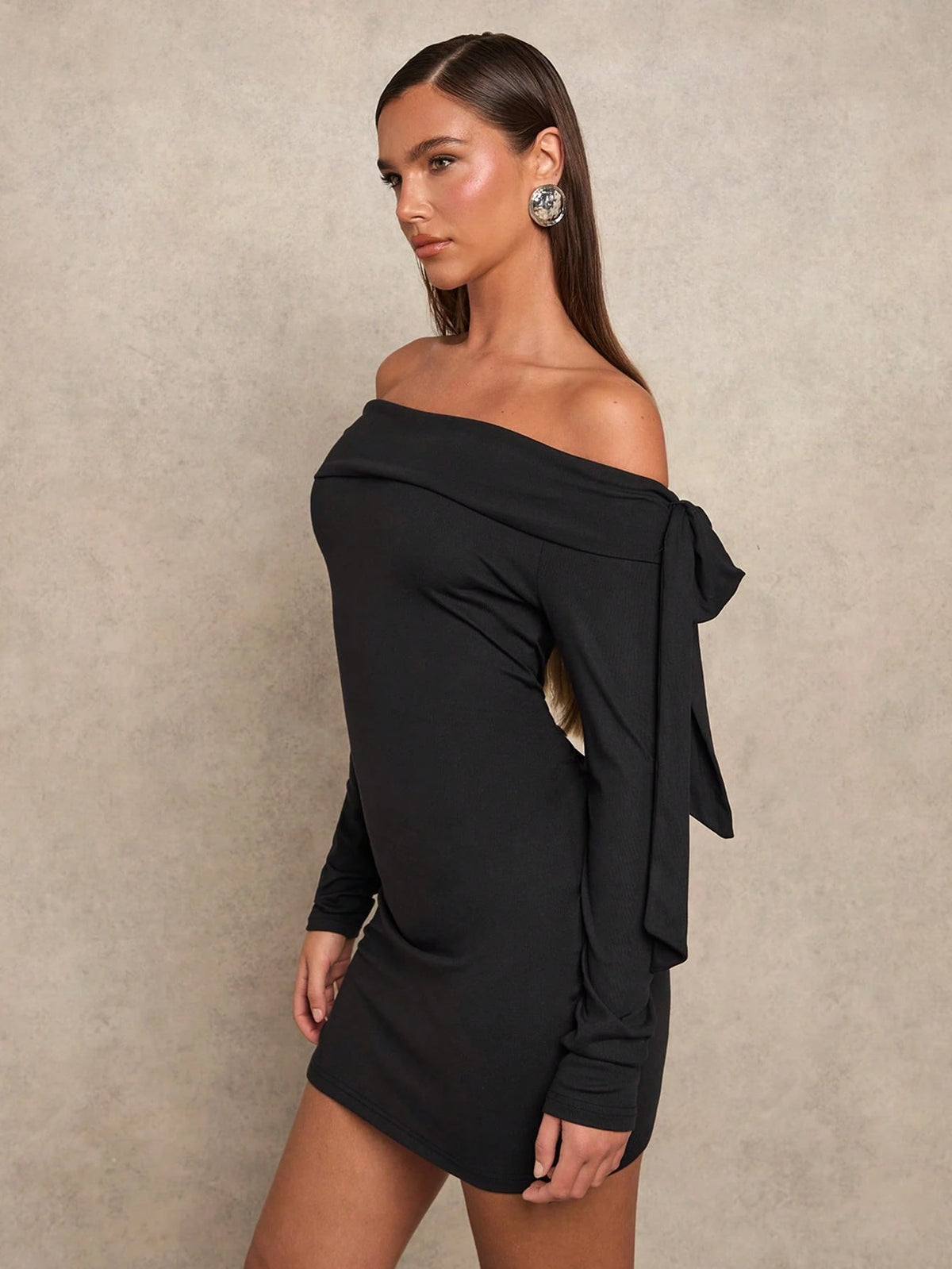 A woman modeling a black off-the-shoulder long-sleeve mini dress with a side bow detail, posed against a textured beige studio backdrop.