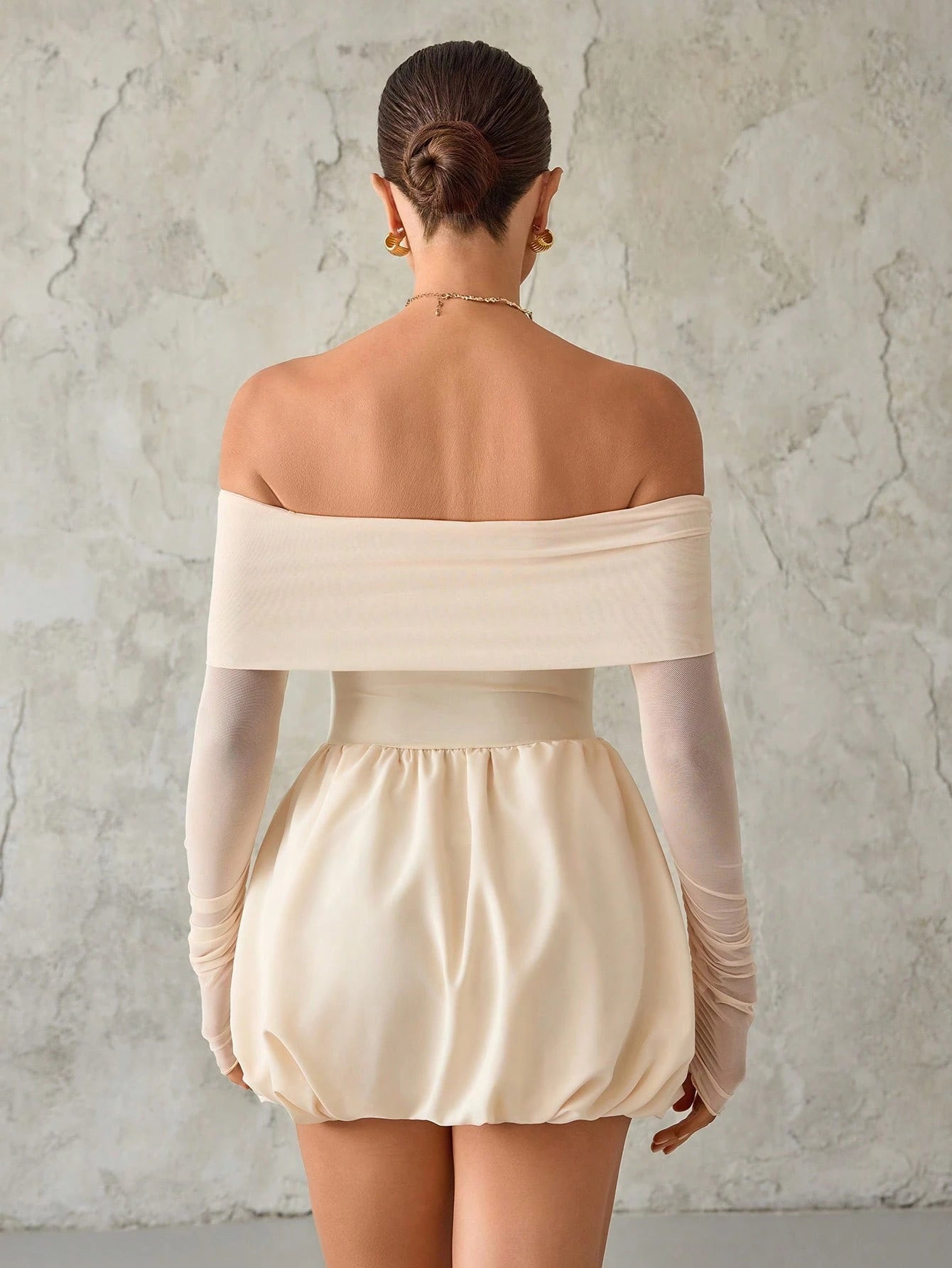 Woman wearing a beige off-shoulder top and shorts against a textured wall.