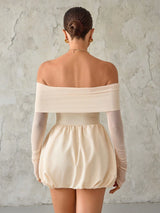 Woman wearing a beige off-shoulder top and shorts against a textured wall.