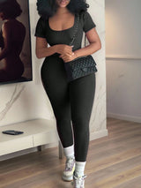 A woman modeling a black ribbed short-sleeve top and matching black ribbed leggings, styled with white socks, white sneakers, and a black quilted shoulder bag with a chain strap in an indoor room setting.