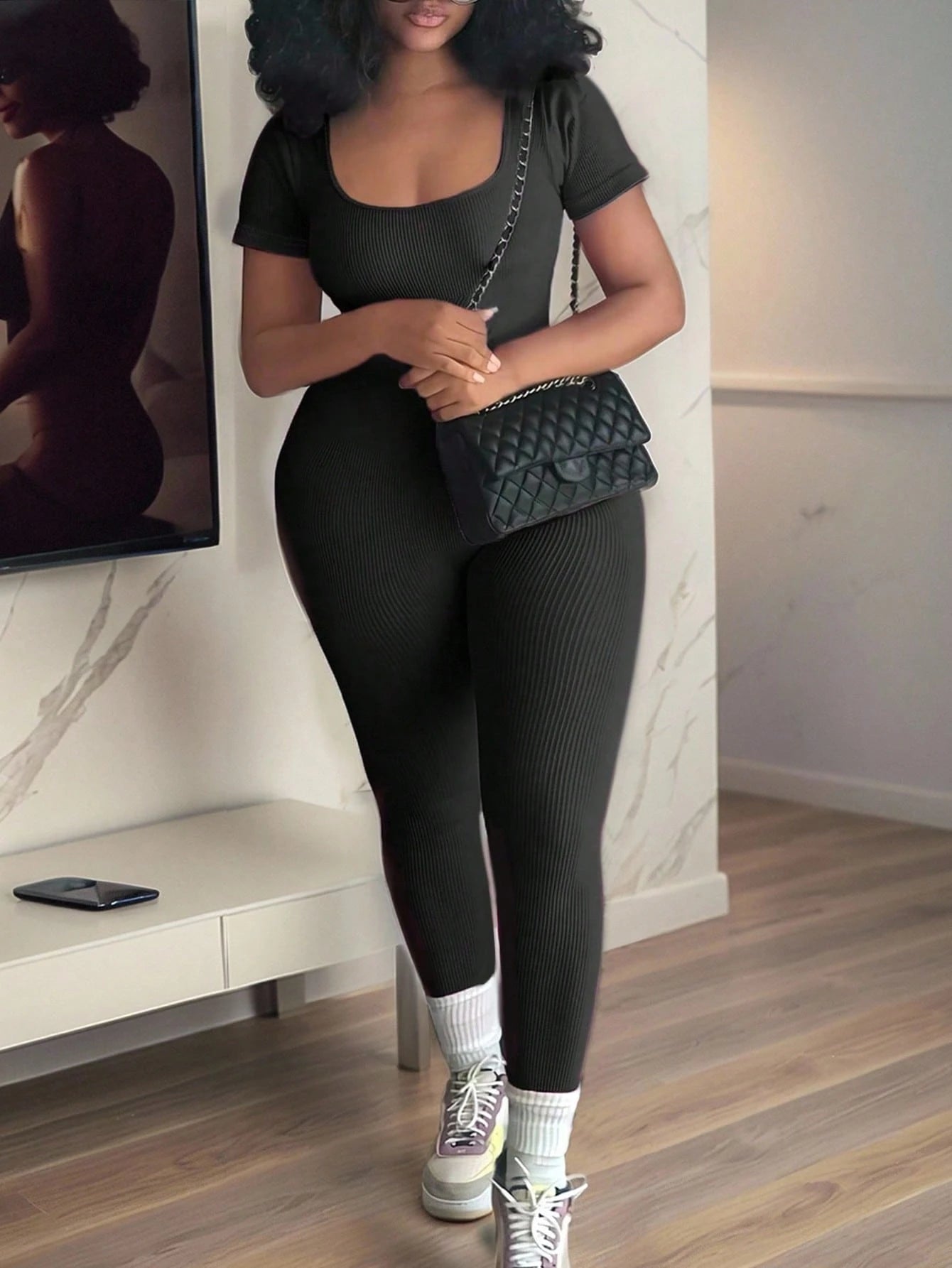 A woman modeling a black ribbed short-sleeve top and matching black ribbed leggings, styled with white socks, white sneakers, and a black quilted shoulder bag with a chain strap in an indoor room setting.