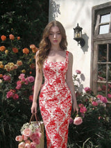 Woman in a red floral dress standing in a garden with flowers and a building in the background