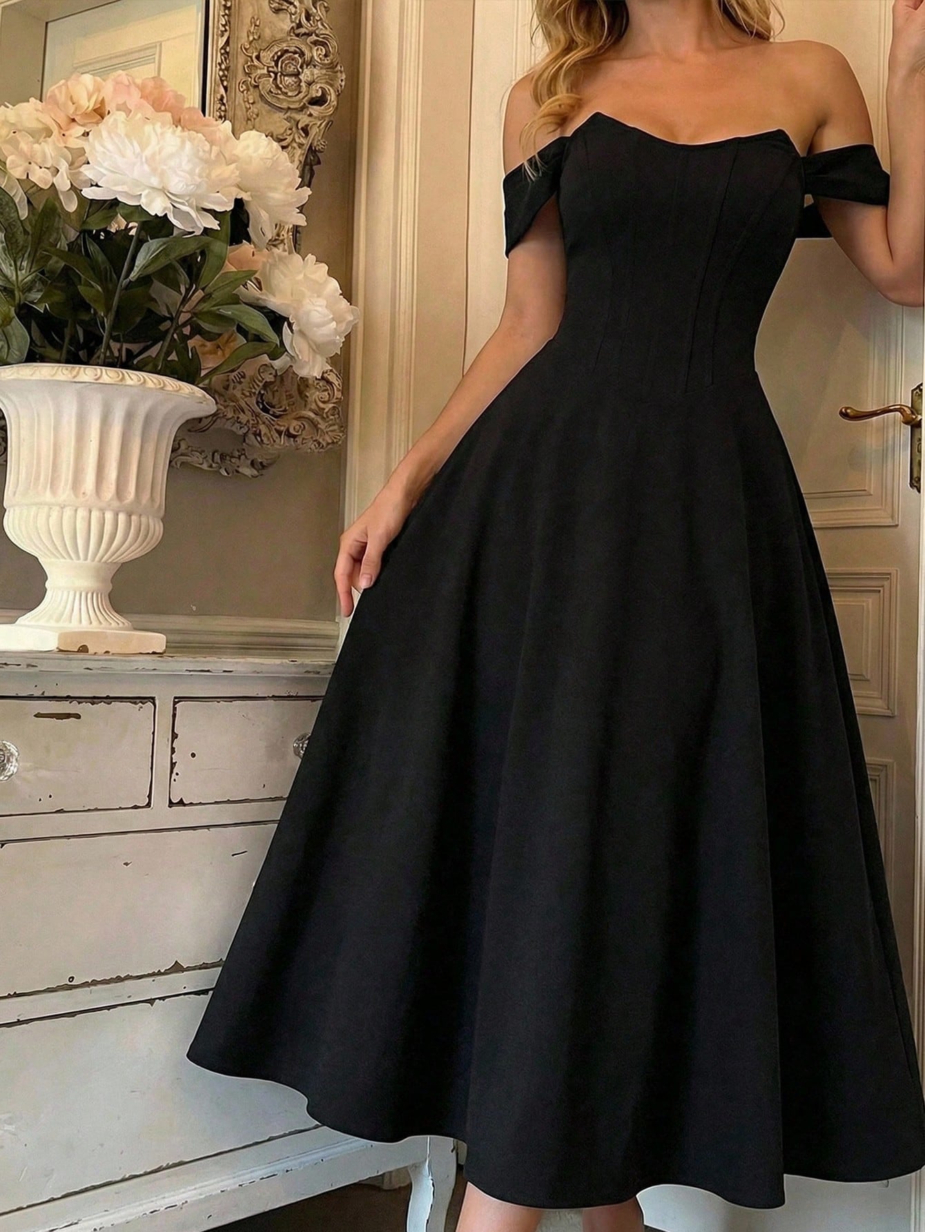 Black off-shoulder dress worn by a person, with a decorative background featuring flowers and a vase.