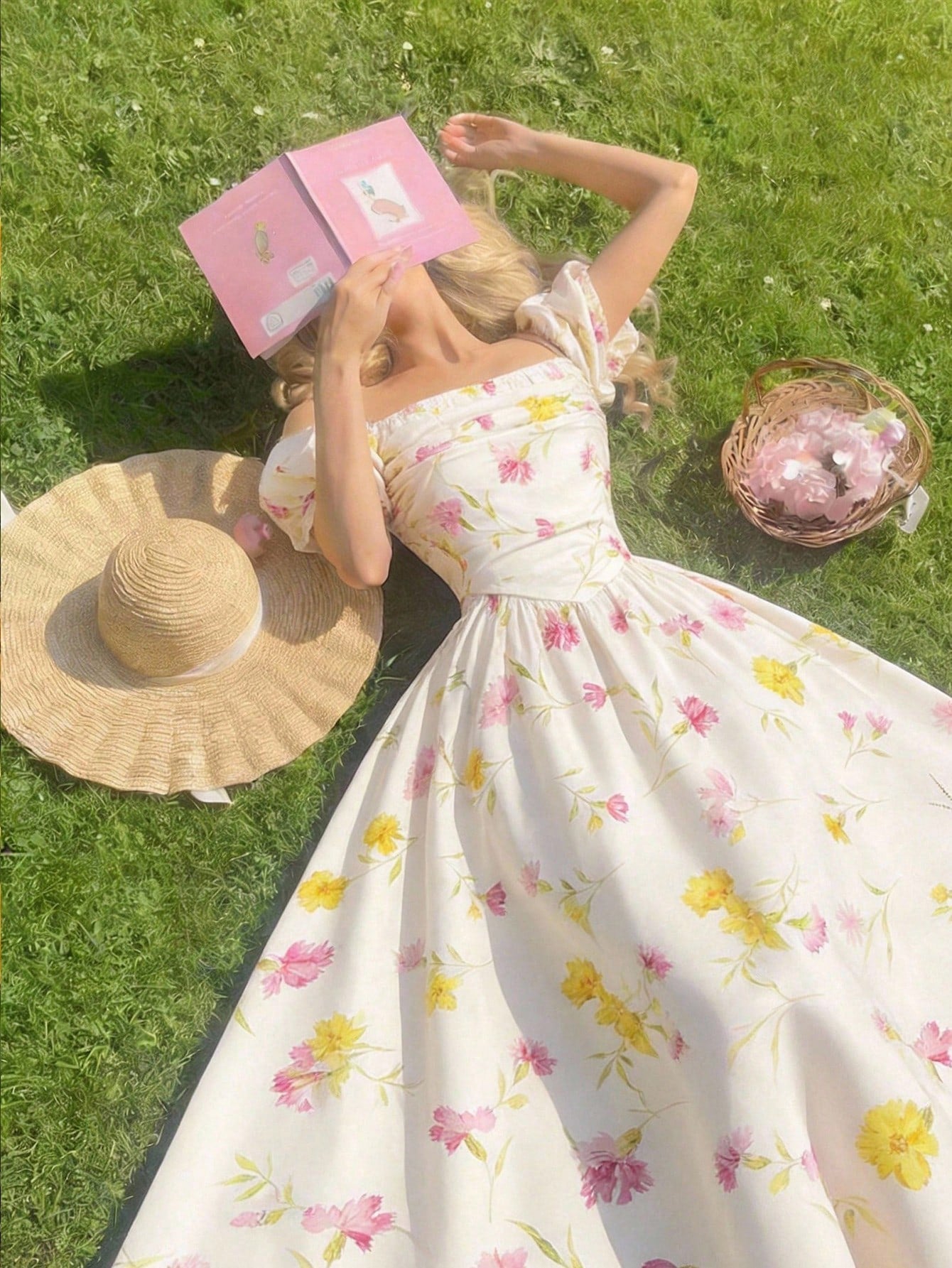 Woman in a floral dress lying on grass with a pink book and straw hat.
