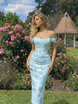 Woman in a light blue floral dress standing in a garden with pink roses and a gazebo.