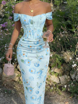 Woman in a light blue floral dress standing outdoors with flowers and greenery around.