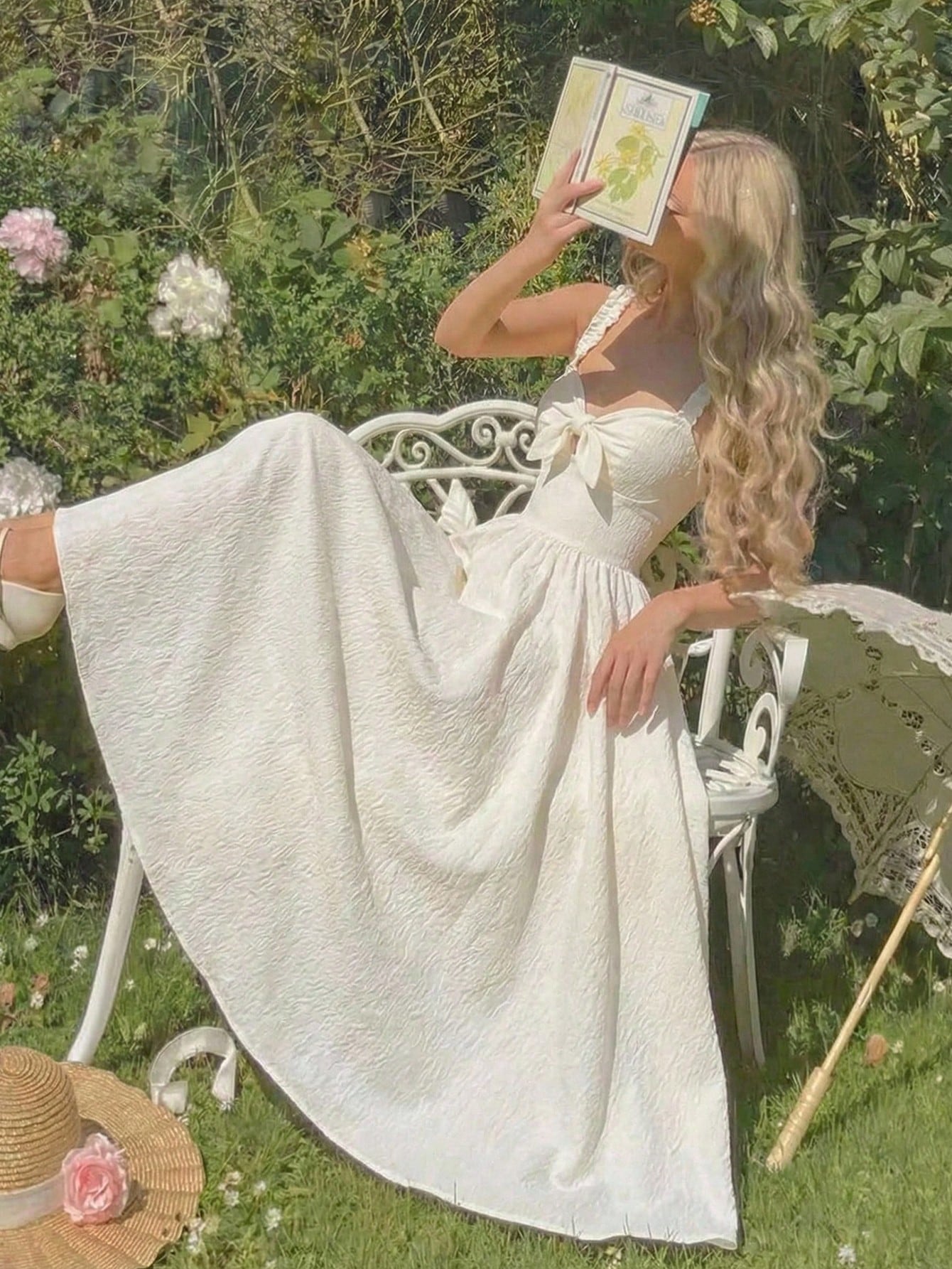 Woman in a white dress sitting on a chair in a garden, holding a book.