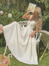 Woman in a white dress sitting on a chair in a garden, holding a book.