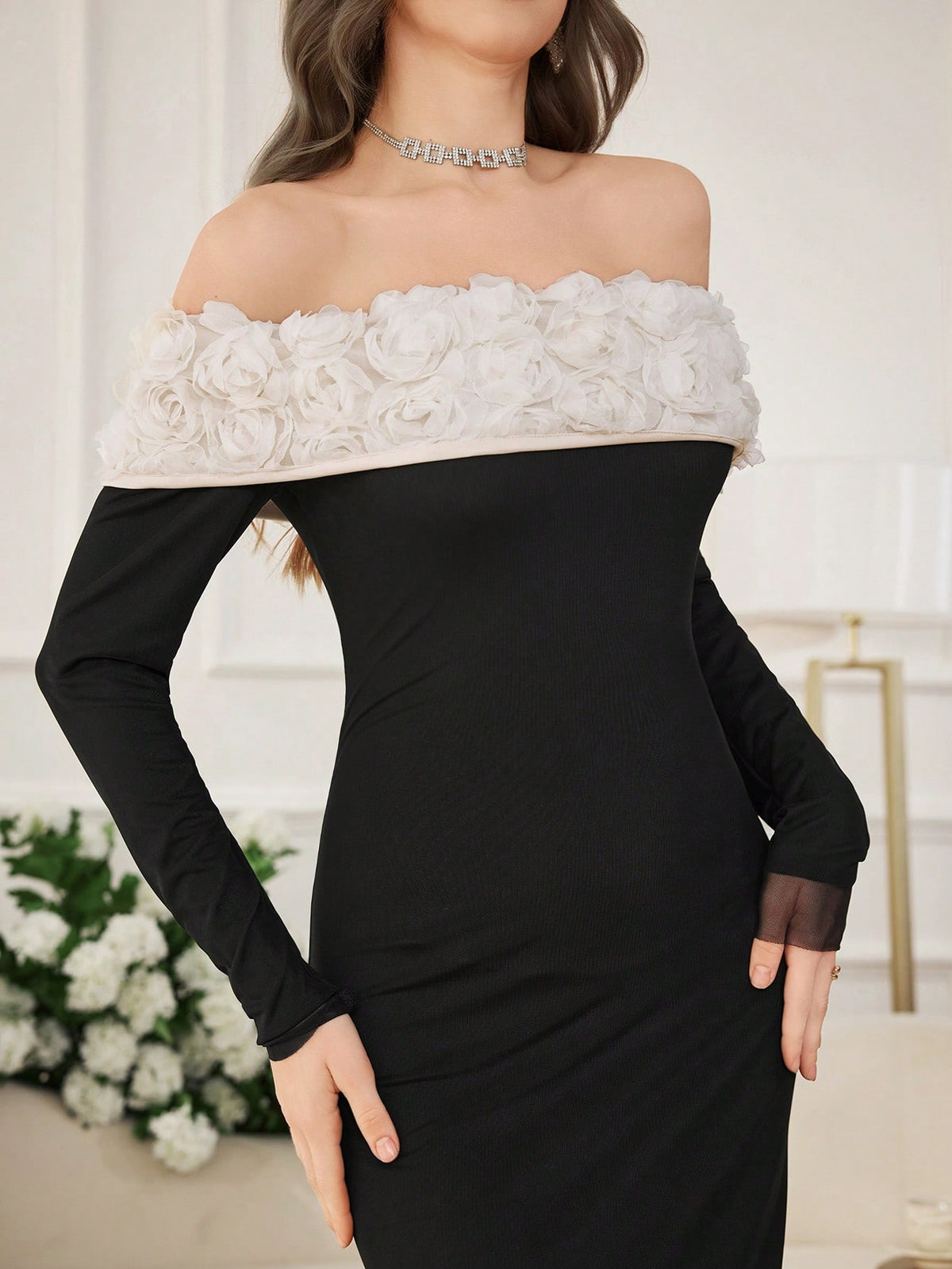 A woman wearing a black off-the-shoulder long-sleeve dress with a wide white floral ruffle trim across the neckline, posed indoors against a light neutral background.