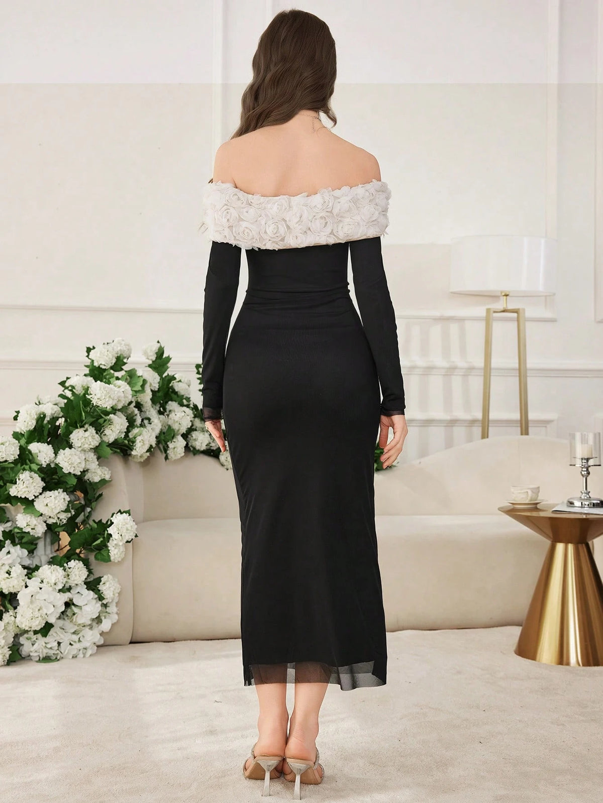 A woman stands with her back to the camera wearing a black off-the-shoulder midi dress with a textured white floral collar, set in a bright, modern living room.