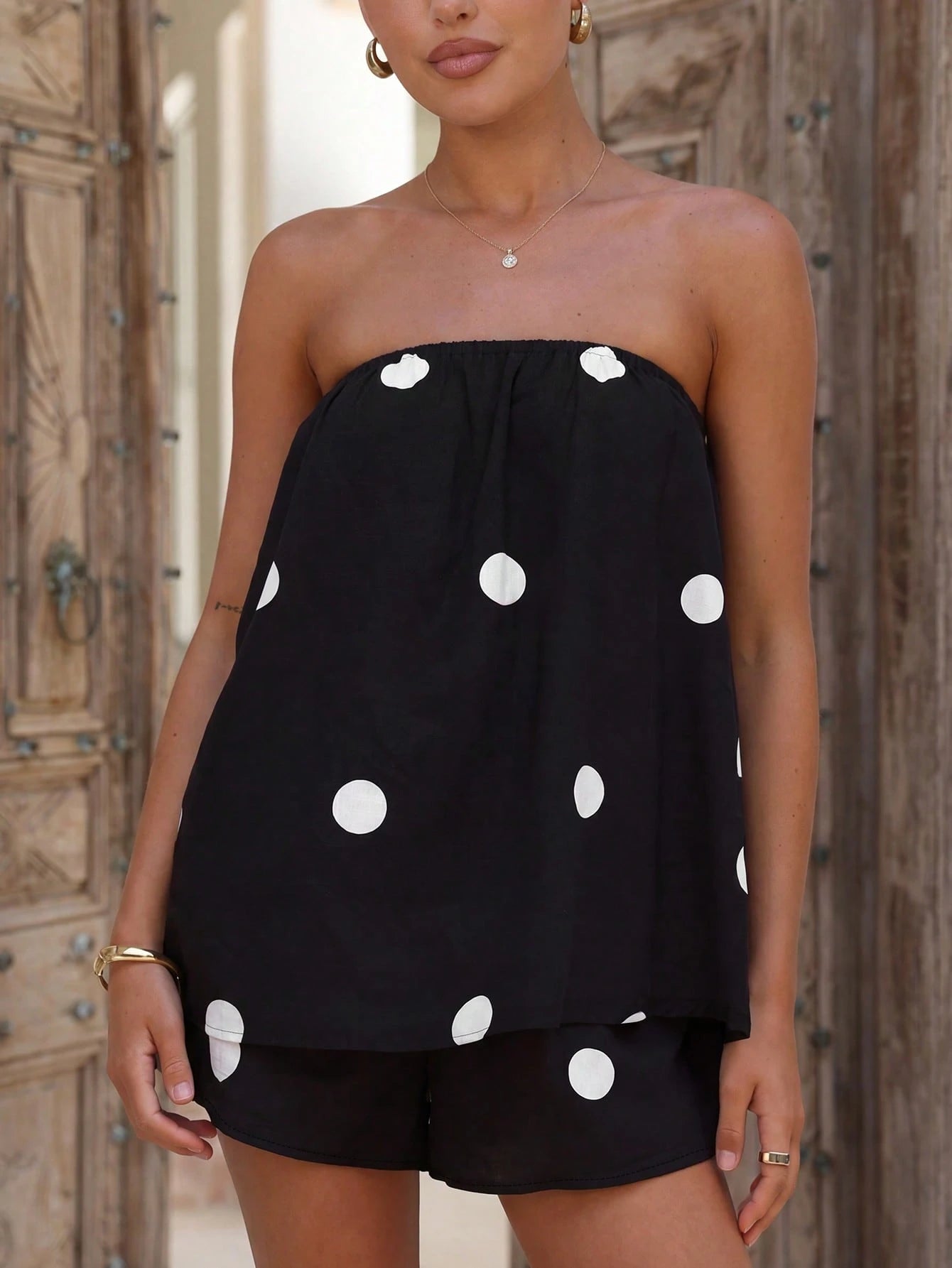 A woman wearing a black strapless top and matching shorts set with large white polka dots, posed in front of rustic wooden doors.