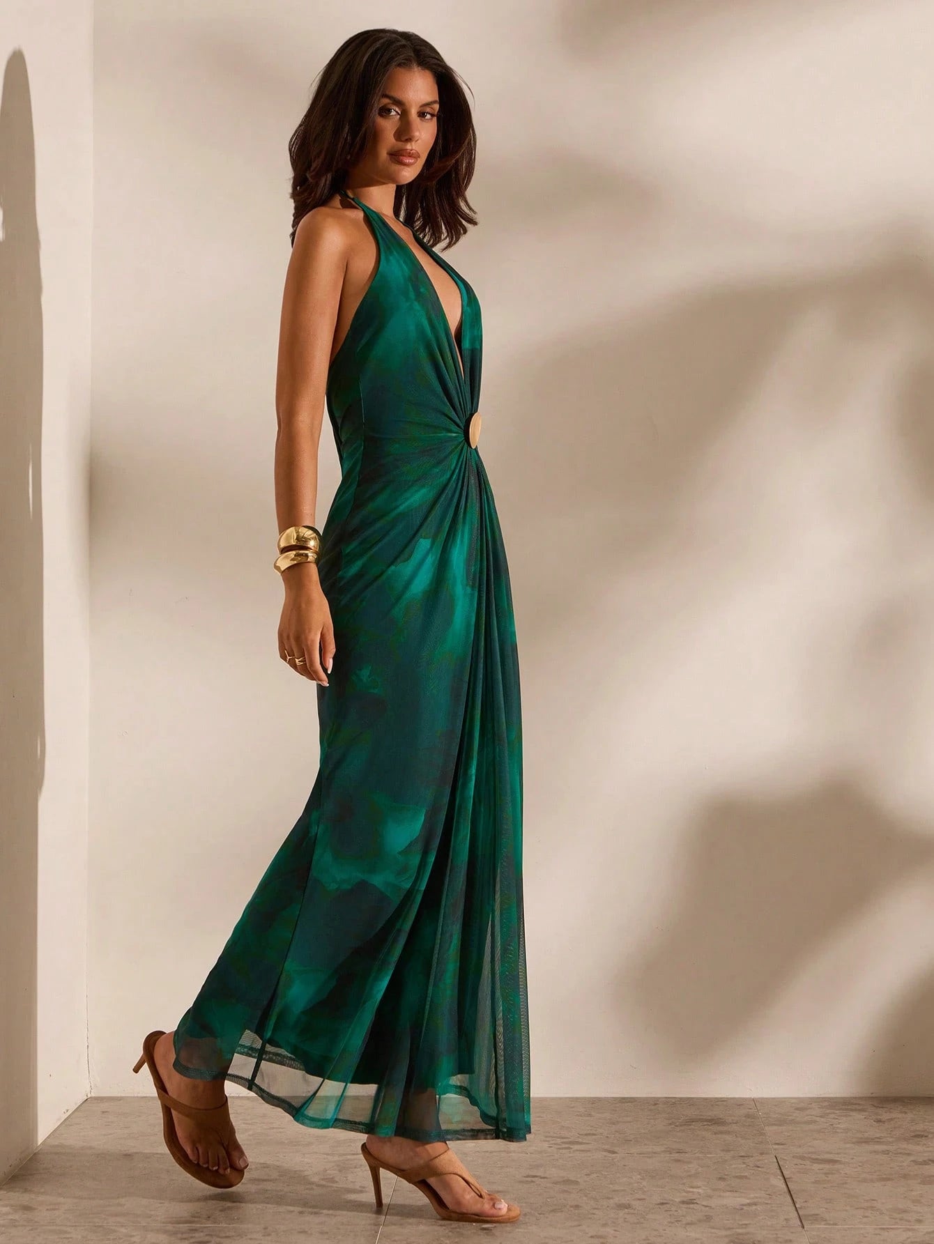 A woman modeling a sleeveless emerald green halter-neck maxi dress with a deep V-neckline and side cutout, styled with brown strappy heels against a warm beige wall with soft shadows.