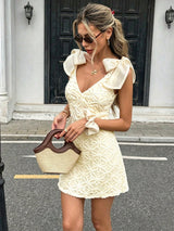 A woman wearing a cream-colored textured mini dress with oversized bow details on the shoulders and waist, holding a woven straw handbag with dark brown handles, standing in front of a building with a black door.