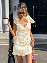 A woman wearing a cream-colored textured mini dress with ruffled cap sleeves, standing outdoors next to a black pole and holding a coffee cup.